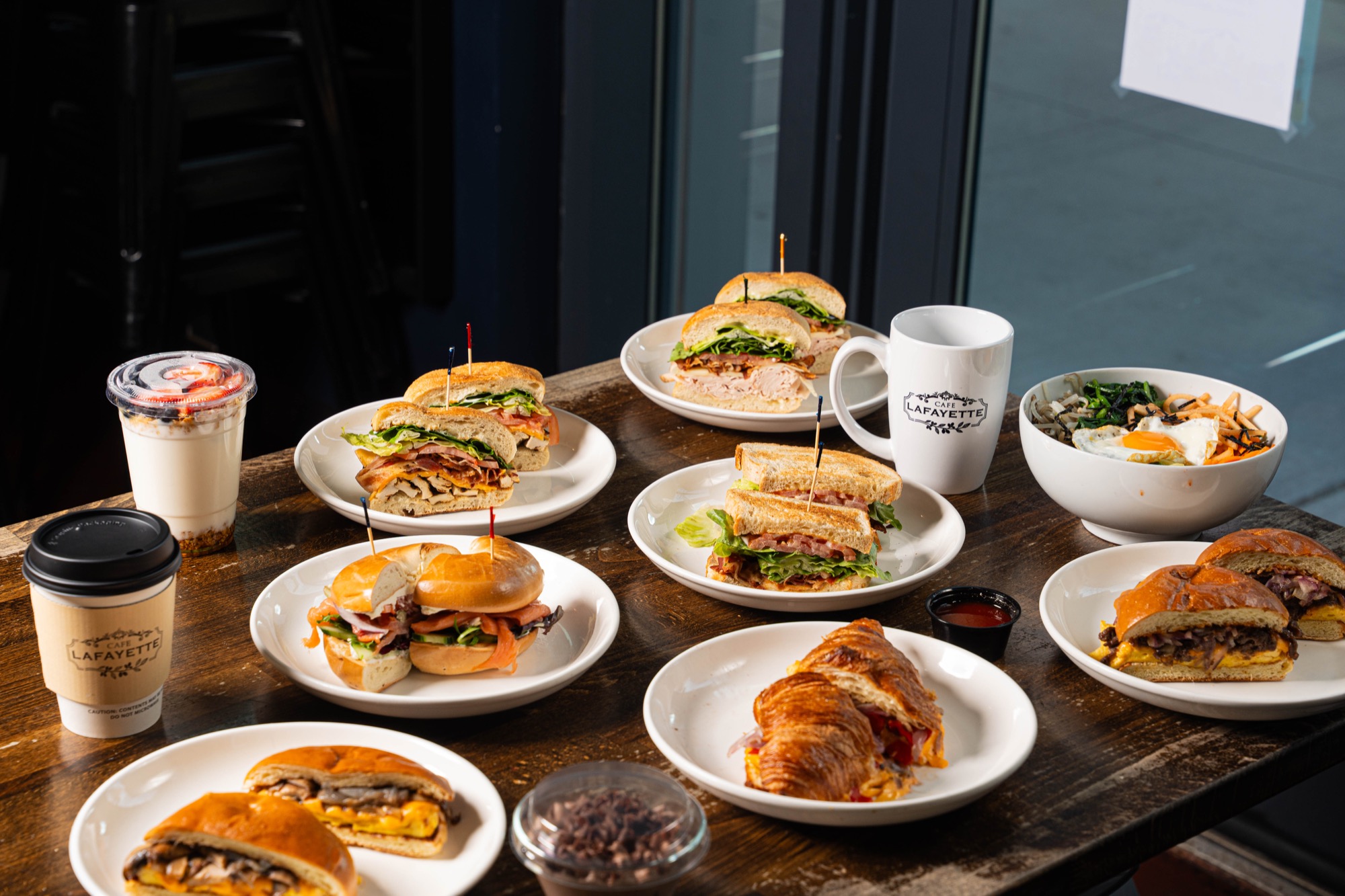 Overhead brunch spread of sandwiches, bowls, and Cafe Lafayette coffee on dark wood