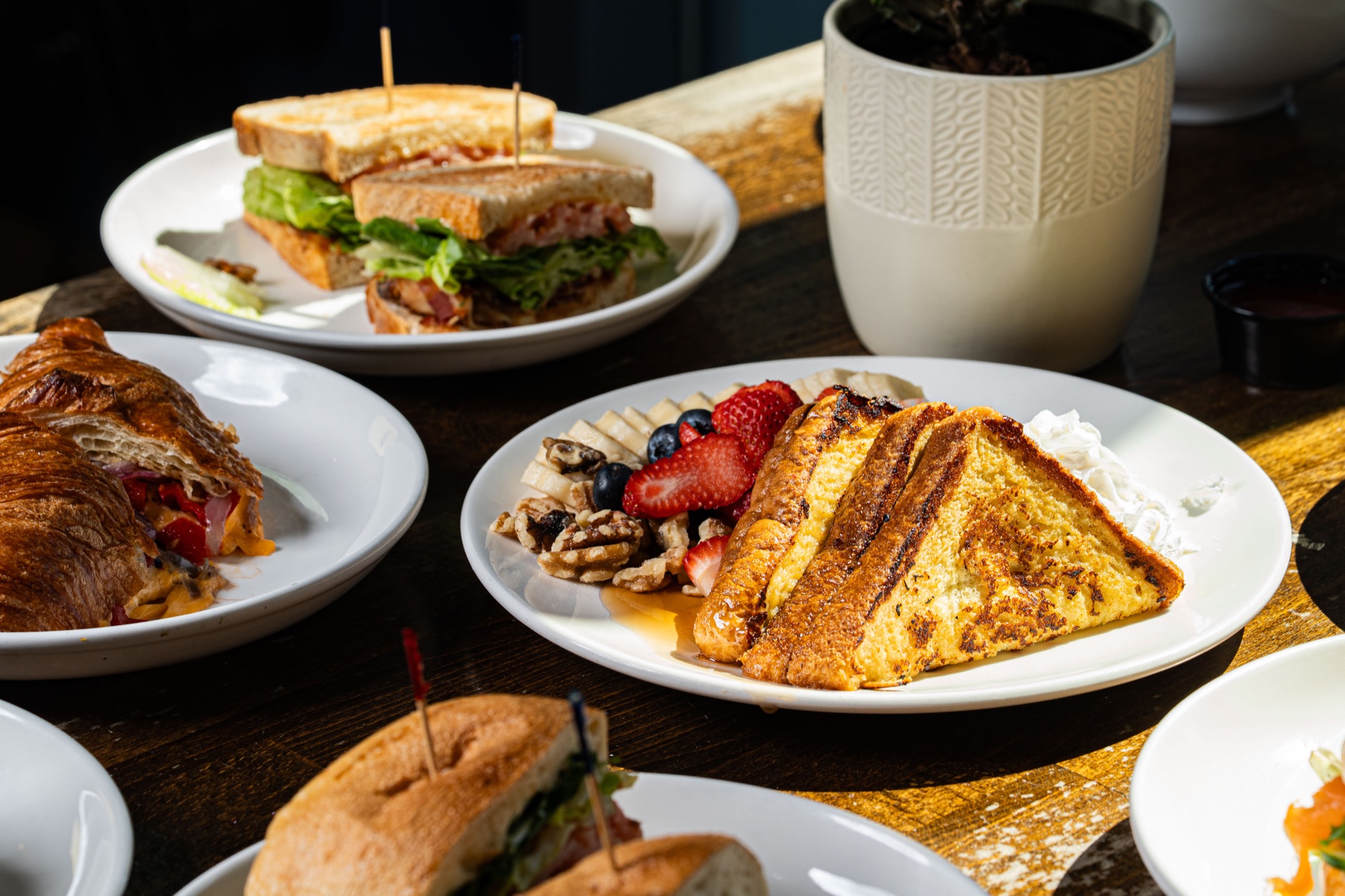 Brunch spread with French toast, berries, croissant sandwich, and club sandwich on dark wood